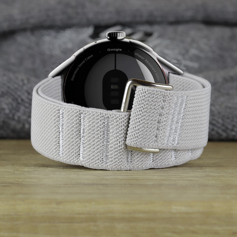 May include: A black smartwatch with a silver band. The watch face is black with white text that reads "Google" and "Water Resistant 5ATM". The back of the watch has the text "Stainless Steel Case".