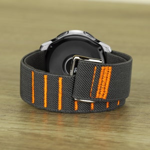 Op de afbeelding: Een grijs en oranje horlogebandje met een zilveren gesp. Het bandje is gemaakt van een zacht, rekbaar materiaal en heeft een sportieve uitstraling.