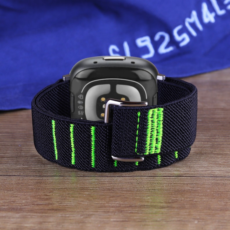 May include: Black and green watch band with a silver buckle. The band is made of a soft, stretchy material and has a textured surface.