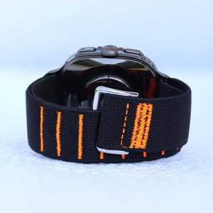 Op de afbeelding: Zwart horlogeband met oranje stiksels. Het band is gemaakt van een rekbaar materiaal en heeft een metalen gesp.