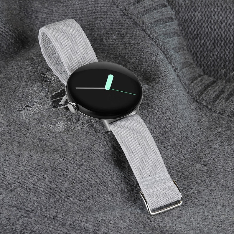 May include: A silver smartwatch with a black face and a gray fabric band. The watch face has a white hour hand and a green minute hand.