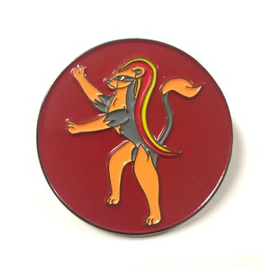 Pode incluir: Um broche de esmalte redondo vermelho com um leão de desenho animado com uma juba amarela e vermelha. O leão está em pé sobre suas patas traseiras com as patas dianteiras levantadas.