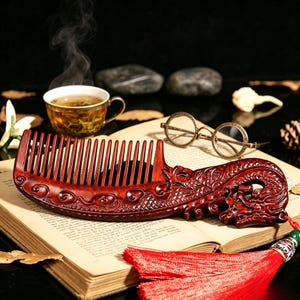 May include: A carved wooden comb in reddish-brown, featuring a dragon design. The comb has fine teeth and a dragon head at the handle. A red tassel is attached. The comb is placed on an open book, with a cup of tea and glasses nearby.