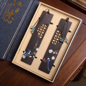 Puede incluir: Dos marcapáginas de madera oscura con intrincados diseños florales y de pájaros, presentados en una caja de regalo. Cada marcapáginas presenta un recorte decorativo y una borla de cuentas. Los marcapáginas están adornados con ilustraciones coloridas de pájaros y flores.