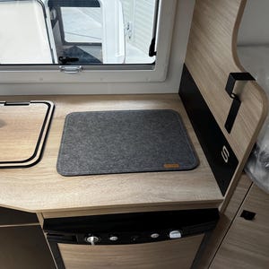 Könnte beinhalten: Innenansicht eines Wohnmobil-Küchenbereichs. Merkmale sind eine helle Holz-Arbeitsplatte mit einer grauen Filzmatte und einem Holzschneidebrett. Ein schwarzer Kühlschrank ist darunter sichtbar.