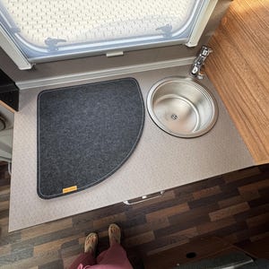 Puede incluir: Vista interior de una cocina de autocaravana. Un fregadero de acero inoxidable junto a una alfombra de fieltro gris oscuro. Una ventana está encima del fregadero. La encimera es gris claro con un mueble de imitación madera a la derecha. El suelo es laminado con aspecto de madera.