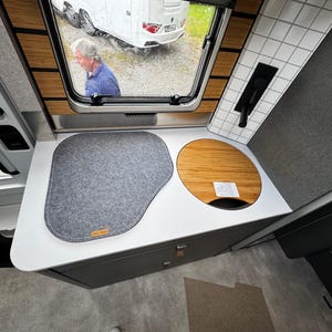 Könnte beinhalten: Innenansicht einer Wohnmobilküche mit grauer Filzmatte und rundem Holzbrett. Die weiße Arbeitsplatte hat einen grauen Schrank darunter. Durch das Fenster ist eine Person zu sehen. Bambusakzente sind vorhanden.