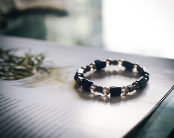 May include: A beaded bracelet featuring a mix of dark square and round beads. The bracelet is primarily black with accents of clear and gold-toned beads. The bracelet is resting on a printed page.