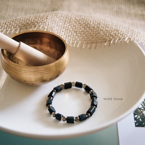 Könnte beinhalten: Ein Armband aus schwarzen Edelsteinen mit silbernen Perlen auf einem weißen Teller neben einer Messing-Singenschale und einem Holzschlägel. Der Teller liegt auf einem Stück Jutegewebe. Der Text "NUDE Stone" ist auf dem Teller sichtbar.