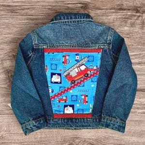 May include: A blue denim jacket with a colorful patch featuring a red fire truck, ambulances, and ladders on a blue background. The patch is bordered with red trim. A children's jacket.
