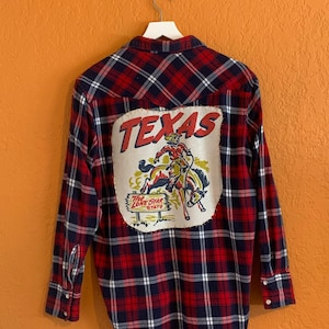 May include: Red and blue plaid flannel shirt with a large patch on the back that says "TEXAS" with a cowboy riding a bucking horse and a sign that says "The Lone Star State".