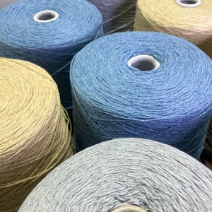 May include: A collection of cylindrical yarn spools in various colors including light blue, beige, and light gray. Each spool features a central white core and is tightly wound with textured thread, arranged in a cluster to show multiple units.