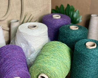 May include: Multiple large cones of yarn in various colors including purple, cream, dark green, and lime green. Each cone is wrapped in a textured, speckled thread. The yarn cones are arranged in a group, with some partially wrapped in plastic.