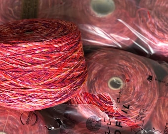 May include: Close-up of several spools of yarn in shades of red, pink, and orange. The yarn is tightly wound, with a visible texture. The spools are partially visible through clear plastic packaging, suggesting a craft supply.
