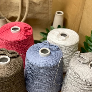 Puede incluir: Cinco grandes bobinas cónicas de hilo dispuestas en grupo. Las bobinas presentan hilo en colores sólidos que incluyen rosa vibrante, azul bígaro suave, blanco nítido, gris claro y un marrón tierra apagado, cada una enrollada en un núcleo blanco.