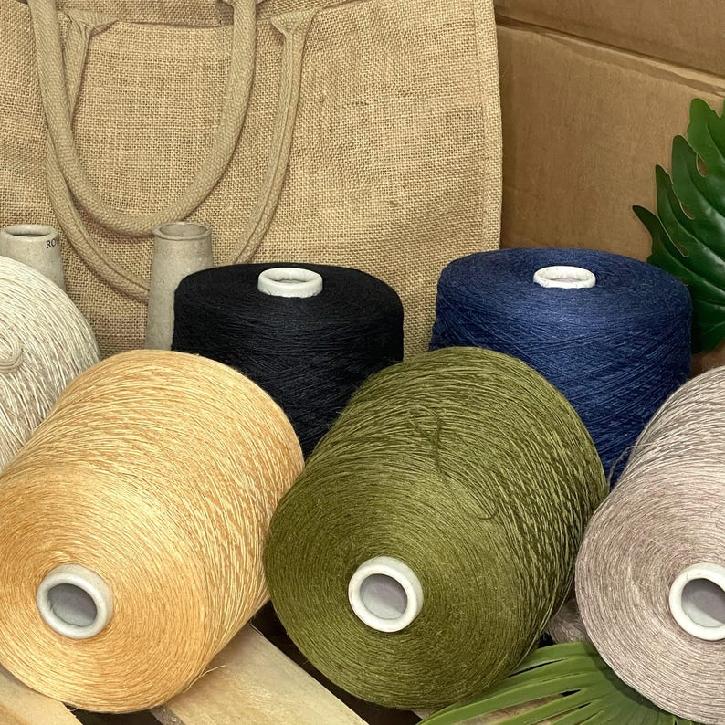 May include: A collection of large yarn cones in various colors including beige, black, olive green, navy blue, and light gray. The cones are arranged in front of a tan burlap tote bag. Each cone features a central white plastic core for winding.