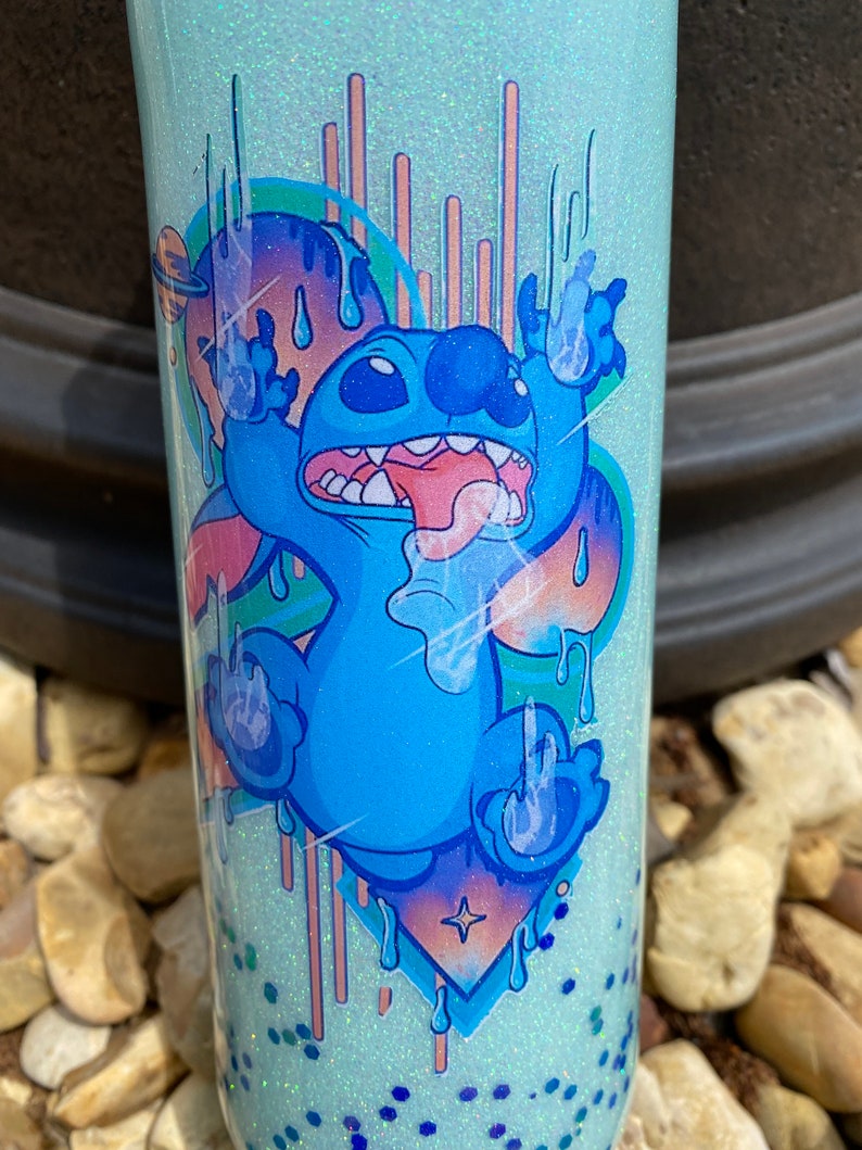 Pu&ograve; includere: Un bicchiere blu chiaro con una finitura scintillante presenta un'immagine di cartone animato di Stitch di Lilo & Stitch. Stitch &egrave; raffigurato con la lingua che sporge e le braccia alzate in un gesto giocoso. Lo sfondo &egrave; blu chiaro con un design astratto colorato.
