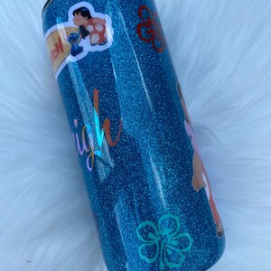 Pu&ograve; includere: Un bicchiere blu con glitter con un designo di fiore di ibisco bianco e un adesivo di Lilo e Stitch. Il bicchiere ha la scritta "Aloha" su di esso.