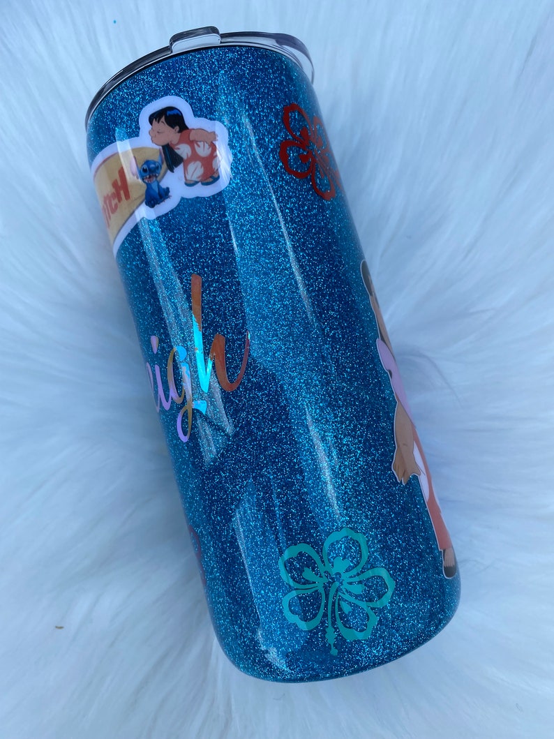 Pu&ograve; includere: Un bicchiere blu con glitter con un designo di fiore di ibisco bianco e un adesivo di Lilo e Stitch. Il bicchiere ha la scritta "Aloha" su di esso.