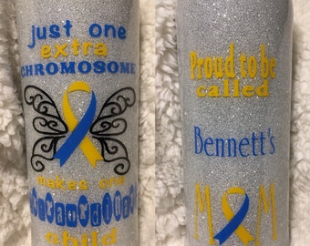 May include: Two silver glitter tumblers with yellow and blue accents. The left tumbler has a butterfly with a yellow and blue ribbon and the text "Just one extra chromosome makes one extraordinary child". The right tumbler has the text "Proud to be called Bennett's Mom" with a yellow and blue ribbon.