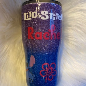 Pu&ograve; includere: Un bicchiere glitter viola e blu con un design bianco e rosso. Il design presenta i personaggi di Lilo e Stitch, un fiore di ibisco e il nome "Rachel".