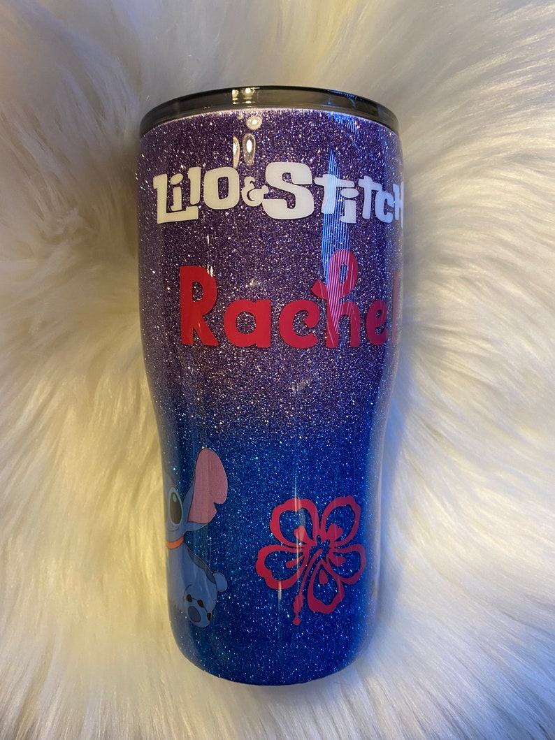 Pu&ograve; includere: Un bicchiere glitter viola e blu con un design bianco e rosso. Il design presenta i personaggi di Lilo e Stitch, un fiore di ibisco e il nome "Rachel".