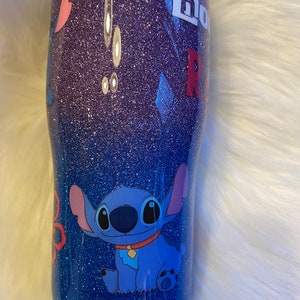 Pu&ograve; includere: Un bicchiere glitter blu e viola con un'immagine di cartone animato di Stitch di Lilo & Stitch. Il bicchiere ha il testo "Lilo" e "Stitch".