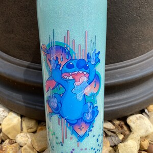 Pu&ograve; includere: Un bicchiere di glitter blu chiaro con un'immagine di cartone animato di Stitch di Lilo & Stitch. Stitch &egrave; blu con un orecchio rosa e gocciola vernice blu e rosa. Lo sfondo &egrave; un glitter blu chiaro.
