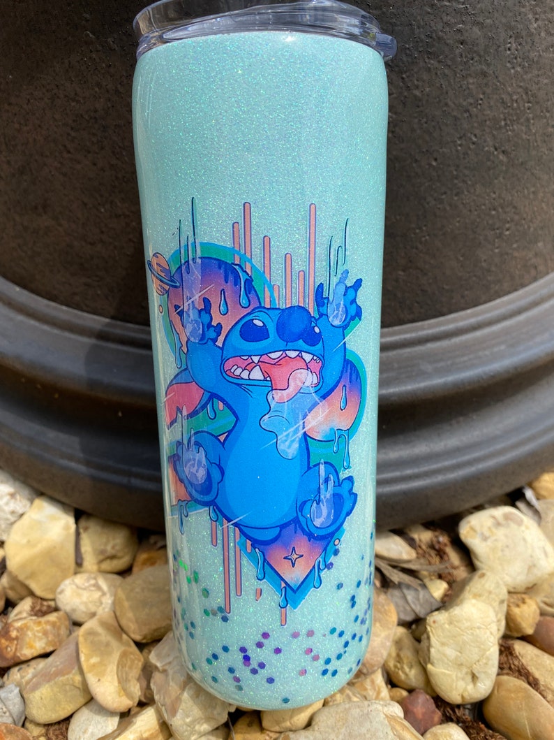Pu&ograve; includere: Un bicchiere di glitter blu chiaro con un'immagine di cartone animato di Stitch di Lilo & Stitch. Stitch &egrave; blu con un orecchio rosa e gocciola vernice blu e rosa. Lo sfondo &egrave; un glitter blu chiaro.