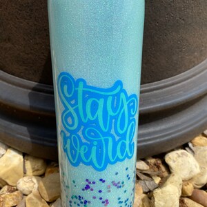 Pu&ograve; includere: Un bicchiere blu chiaro con una finitura scintillante e le parole "Stay Weird" in lettere blu. Il bicchiere &egrave; pieno di glitter blu, verde e viola.