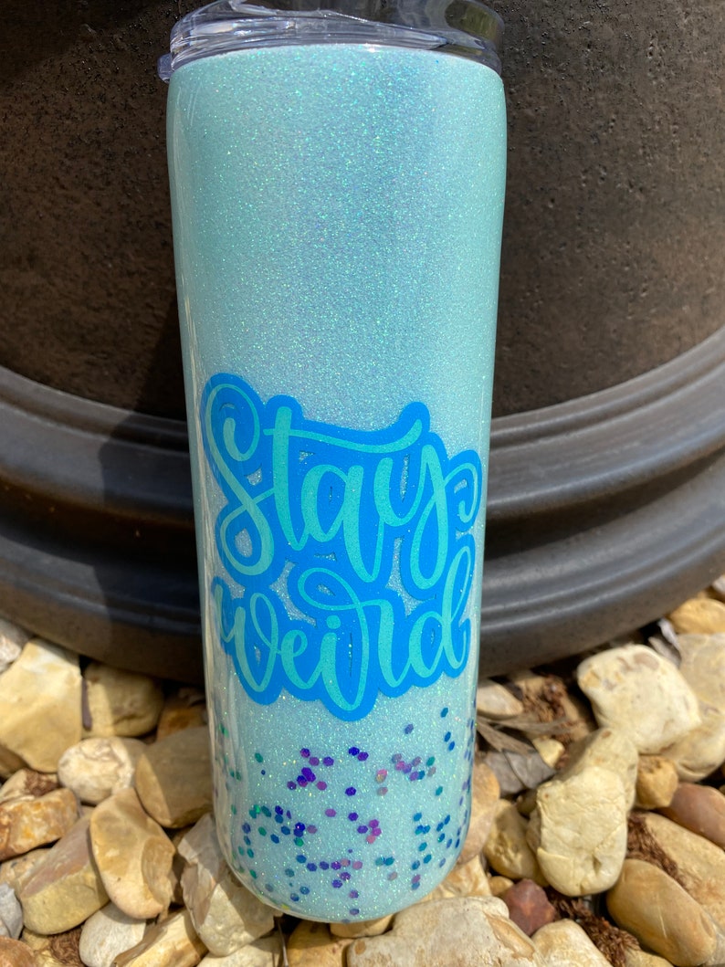 Pu&ograve; includere: Un bicchiere blu chiaro con una finitura scintillante e le parole "Stay Weird" in lettere blu. Il bicchiere &egrave; pieno di glitter blu, verde e viola.