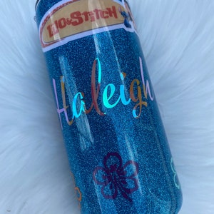 Pu&ograve; includere: Un bicchiere glitter blu turchese con un adesivo di Lilo & Stitch, il nome "Haleigh" in lettere iridescenti e adesivi di fiori viola.