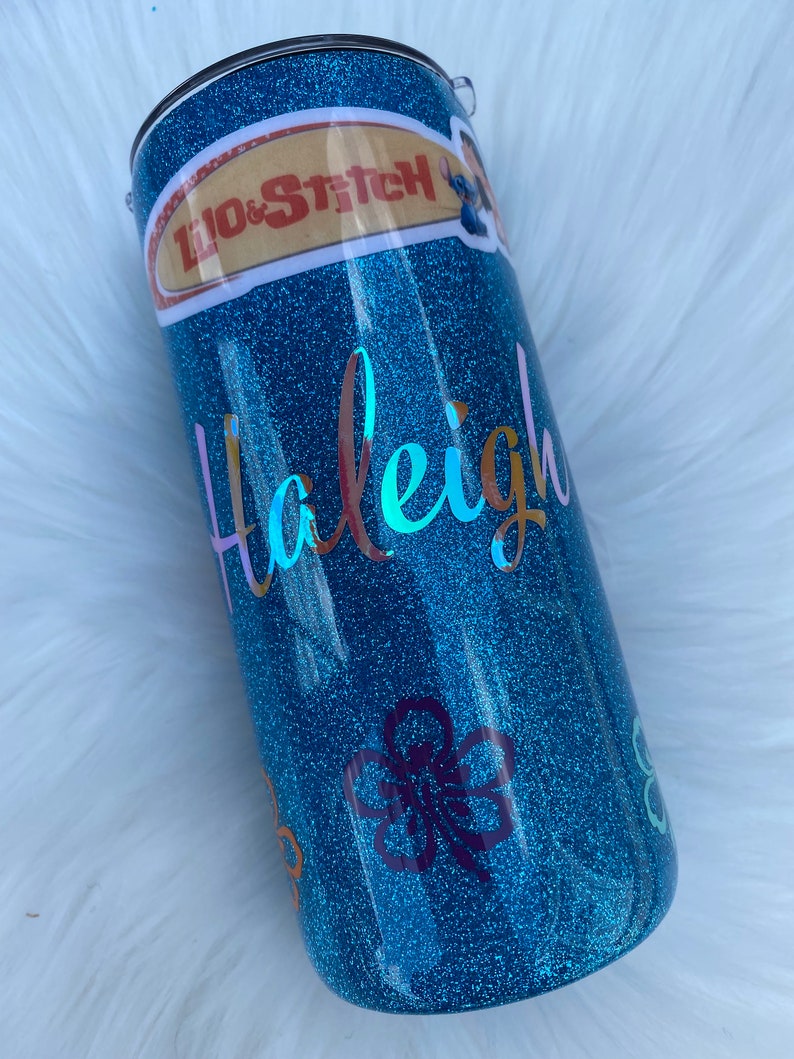 Pu&ograve; includere: Un bicchiere glitter blu turchese con un adesivo di Lilo & Stitch, il nome "Haleigh" in lettere iridescenti e adesivi di fiori viola.
