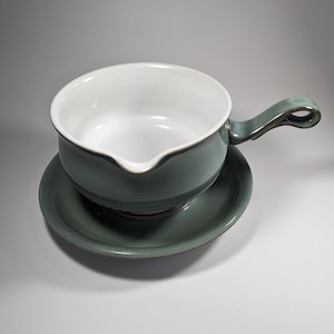 May include: A green ceramic gravy boat with a white interior and a matching saucer. The boat has a handle on the side.