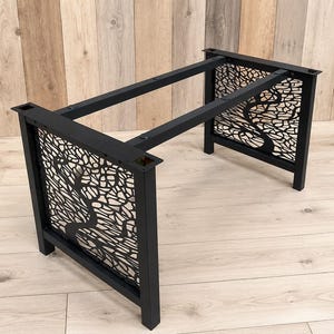 May include: A black metal table base featuring two rectangular end frames with intricate, laser-cut tree branch patterns. The structure includes two horizontal support bars connecting the ends, with flat mounting plates at the top corners.