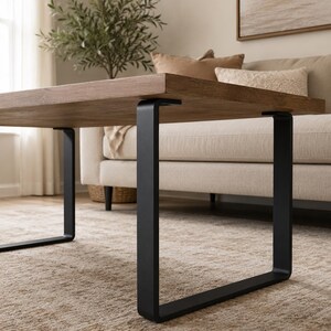 May include: A rectangular coffee table featuring a thick, medium-brown wood grain tabletop supported by two sturdy, matte black U-shaped metal legs. The design showcases a clean, flat surface with visible wood texture and sharp, angular metal supports.