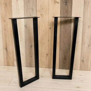 May include: A pair of black metal table legs featuring a trapezoidal frame design. Each leg consists of two vertical bars connected by a horizontal base bar, with a flat mounting plate at the top for attachment to a tabletop surface.