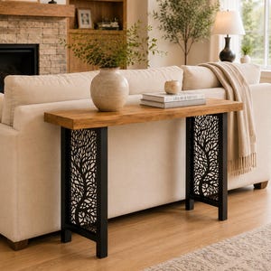 May include: A rectangular console table with a natural wood grain top and two black metal legs featuring an intricate laser-cut tree branch pattern. The table is positioned behind a beige sofa and holds a ceramic vase and two stacked books.