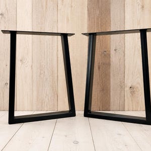 May include: A pair of black metal table legs featuring a trapezoidal frame design. Each leg consists of two vertical supports connected by a horizontal base bar and a flat mounting plate at the top, constructed from solid square metal tubing.