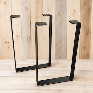 May include: Two U-shaped furniture legs made of matte black metal. Each leg features a flat horizontal base, two vertical sides, and a top mounting plate with two pre-drilled circular holes for attachment to a table or bench surface.