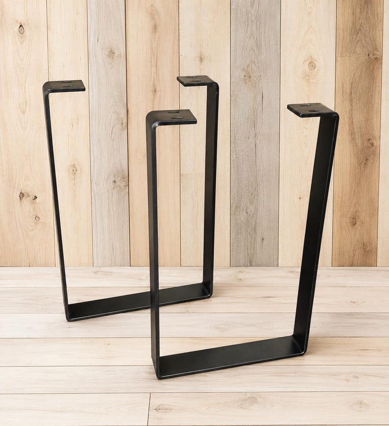 May include: Two U-shaped furniture legs made of matte black metal. Each leg features a flat horizontal base, two vertical sides, and a top mounting plate with two pre-drilled circular holes for attachment to a table or bench surface.