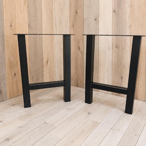 May include: A pair of black metal table legs featuring a trapezoidal frame design. Each leg consists of two vertical supports connected by a horizontal crossbar at the base, with a flat mounting plate at the top for attachment to a tabletop surface.