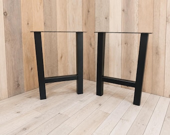 May include: A pair of black metal table legs featuring a trapezoidal frame design. Each leg consists of two vertical supports connected by a horizontal crossbar at the base, with a flat mounting plate at the top for attachment to a tabletop surface.