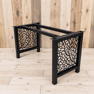 May include: A black metal table base featuring two rectangular side panels with an intricate, laser-cut organic leaf or branch pattern. The frame consists of four vertical legs connected by horizontal support bars with pre-drilled mounting holes.