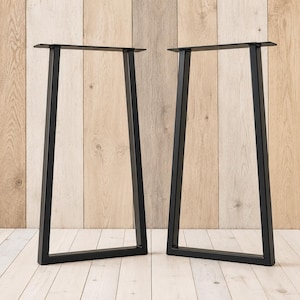 May include: Two black metal table legs with a trapezoidal frame design. Each leg features a flat rectangular mounting plate at the top, two angled vertical supports, and a horizontal base bar, constructed from square-profile metal tubing.