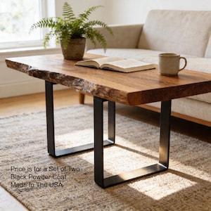 May include: A rectangular wooden coffee table with a live edge top and black metal legs. The table is made of natural wood and has a warm, brown finish. An open book and a mug sit on the table. A potted plant is also visible.