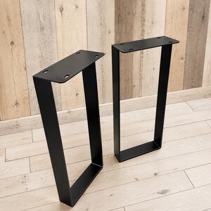 May include: A pair of matte black metal table legs with a U-shaped design. Each leg features a flat rectangular mounting plate at the top with four pre-drilled circular holes for attachment, constructed from solid flat bar steel.