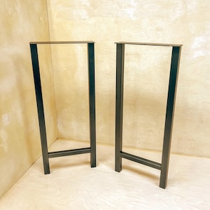 May include: Two black metal table legs with a rectangular top and a single crossbar. The legs are standing on a light brown surface.
