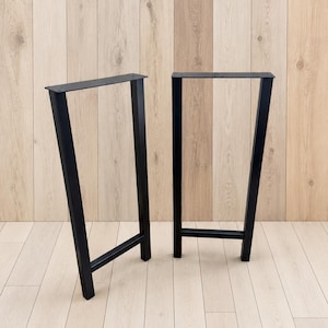 May include: Two black metal table legs with a trapezoidal frame design. Each leg features a flat rectangular mounting plate at the top with pre-drilled holes for attachment and a horizontal support bar connecting the two vertical posts.