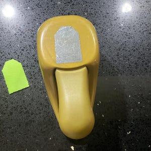 May include: A mustard yellow paper punch tool with a rounded top and a lever mechanism. The tool features a silver-colored metal cutting area at the top and is positioned next to a single, small, light green paper tag with a pointed top edge.